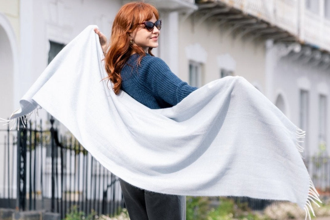Woman with red hair holding the Smoke oversized blanket scarf, showcasing its herringbone weave and tassels, on a street with townhouse-style buildings.