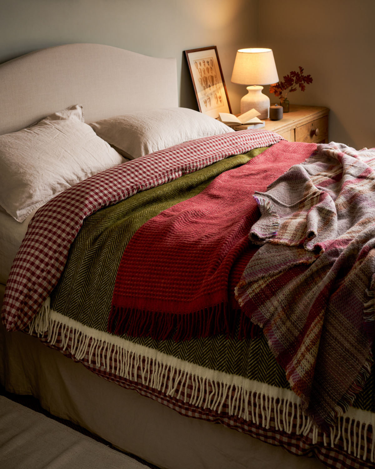 A cosy bedroom scene with a bed dressed in layered woven throw blankets in red, olive green, beige, and plaid, with a matching headboard and bedside lamp in warm lighting.