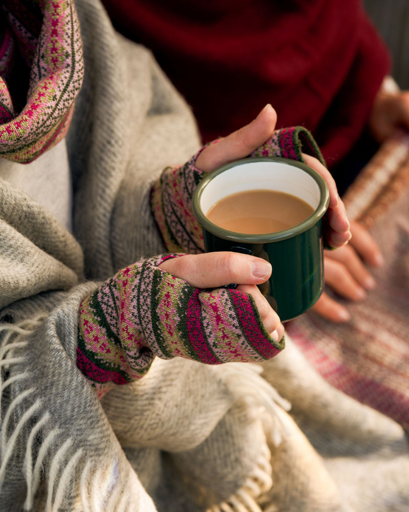 A close-up lifestyle shot of a person wrapped in a cozy beige knit blanket and matching scarf, wearing patterned fingerless knit gloves while holding a dark green enamel mug filled with a hot beverage.