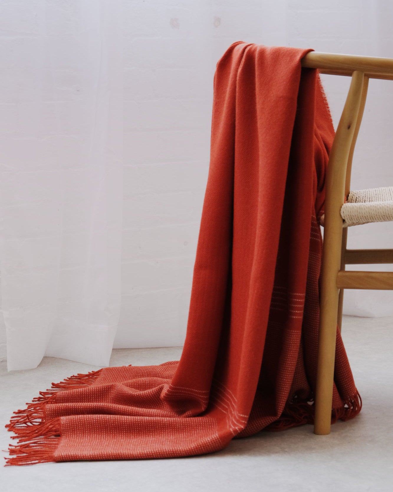 Alpaca Blanket Gift Box: Flame Orange featuring a rust-red woven alpaca blanket with fringe, draped over a light wood chair, set in a softly lit room.