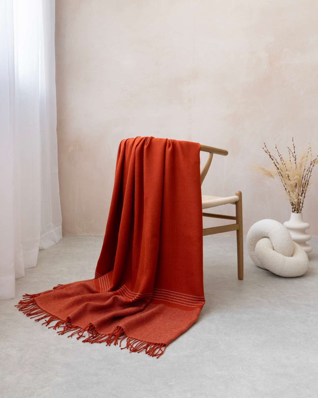 Alpaca Blanket Gift Box: Flame Orange features a rust-red throw on a woven chair, paired with a white vase and knot cushion for minimalist elegance.