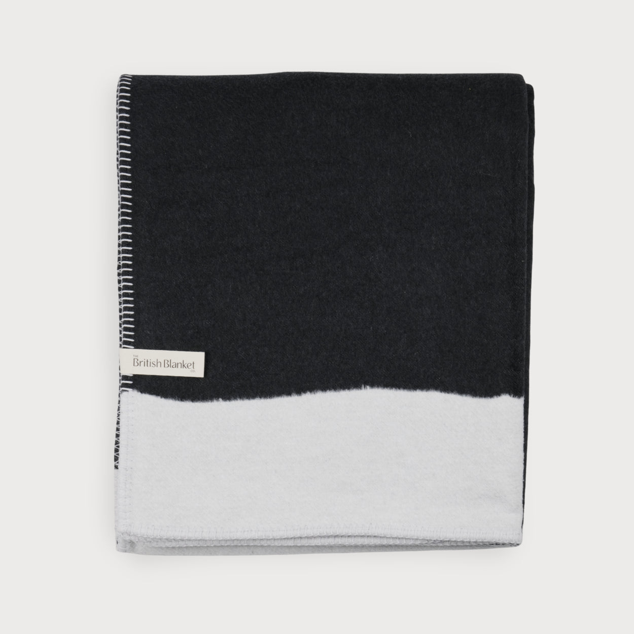 Cloud Cotton Blanket: Strata in Flint, featuring a two-tone wool design with whip-stitched edges and a label from The British Blanket Co., offering cosy, hypoallergenic comfort.