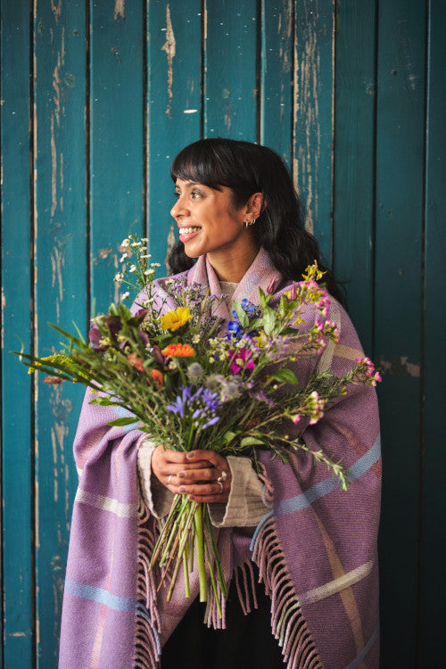 Smiling woman holding a bunch of wild flowers and wrapped in a lavender purple Garden flowers merino wool throw blanket collection by The British Blanket Company online shop