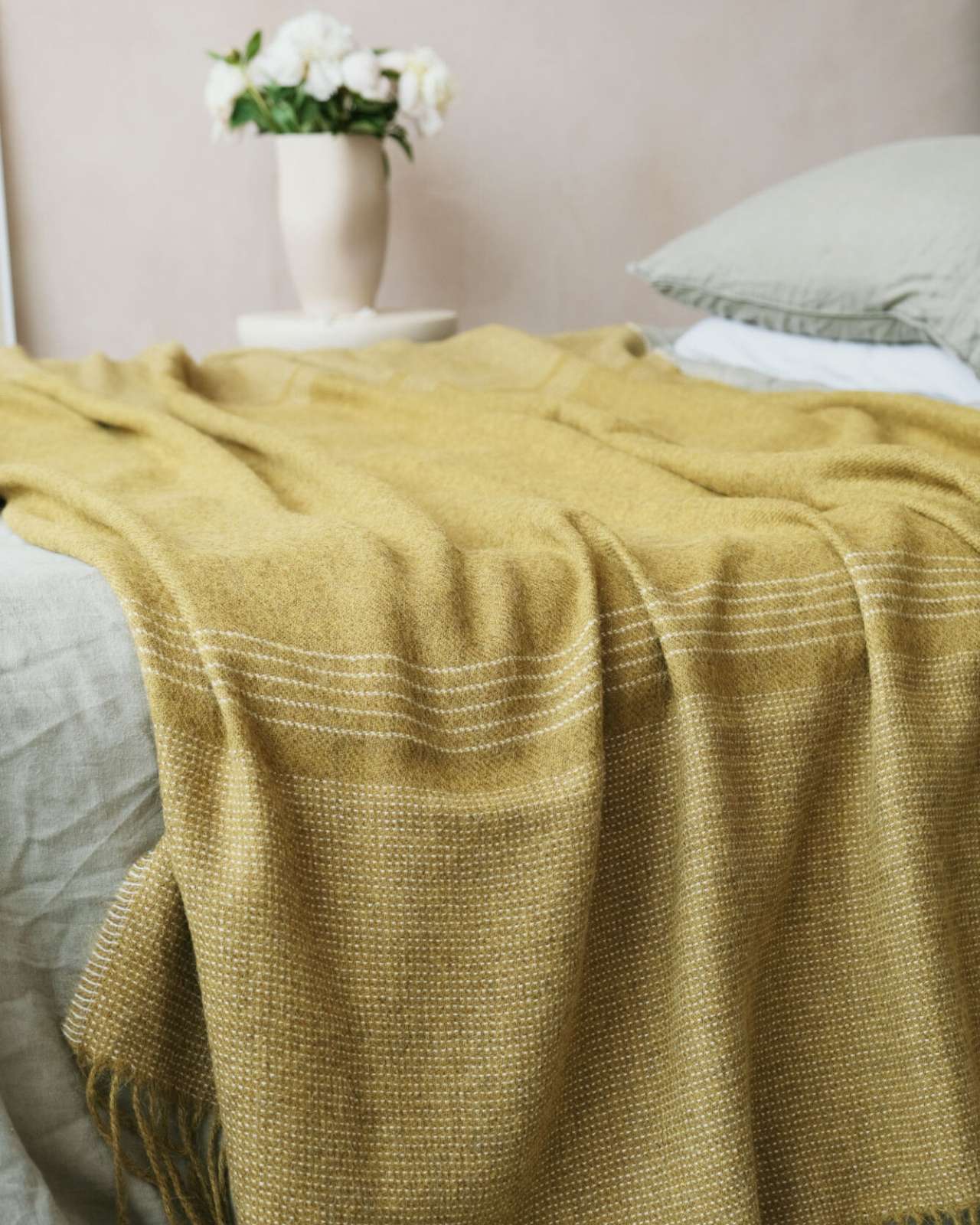 Alpaca Blanket Gift Box: Golden Moss features a mustard-yellow throw blanket elegantly draped on a bed, paired with a vase of white blossoms on a side table.