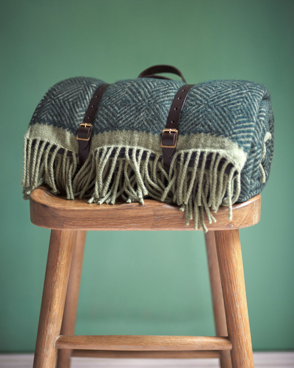 Green wool picnic blanket with leather straps on stool from The British Blanket Company