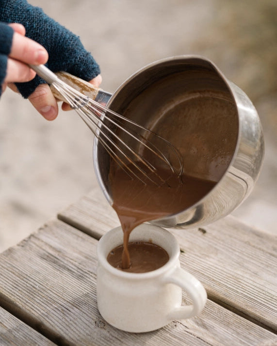 Pouring Artisan Hot Chocolate from a saucepan into a mug, with a whisk inside, set on a rustic wooden surface, suggesting an indulgent, handcrafted experience.
