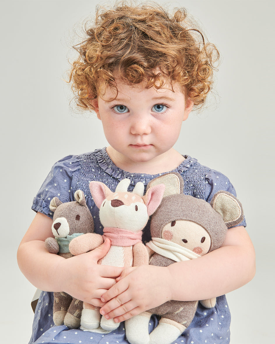 Toddler with curly hair holds a knitted teddy bear soft toy, alongside a deer and mouse plush, wearing a blue polka-dot dress.