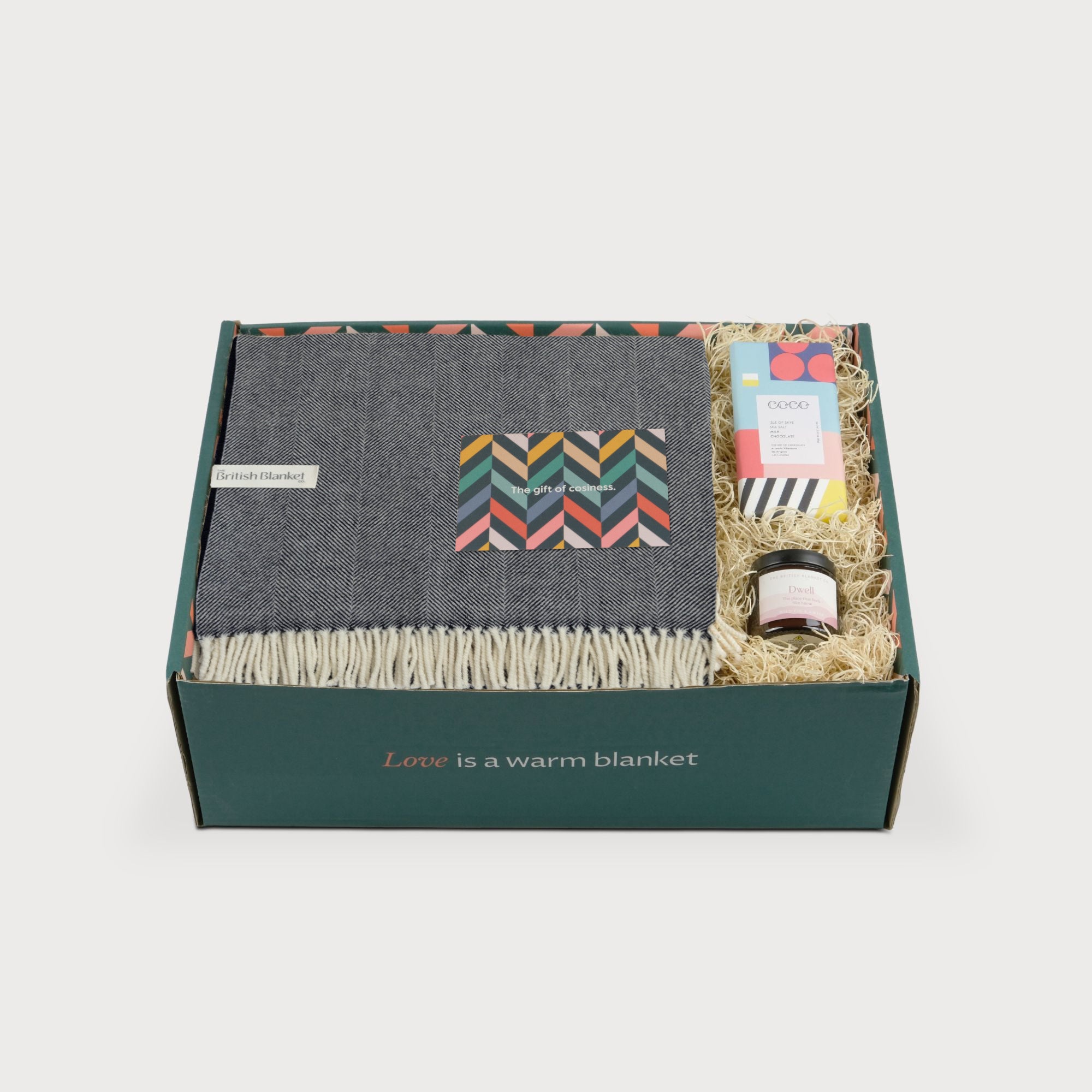 Merino Blanket Gift Box: Navy Blue includes a grey herringbone wool blanket, amber jar candle, COCO chocolate bar, and gift card, all nestled in straw.