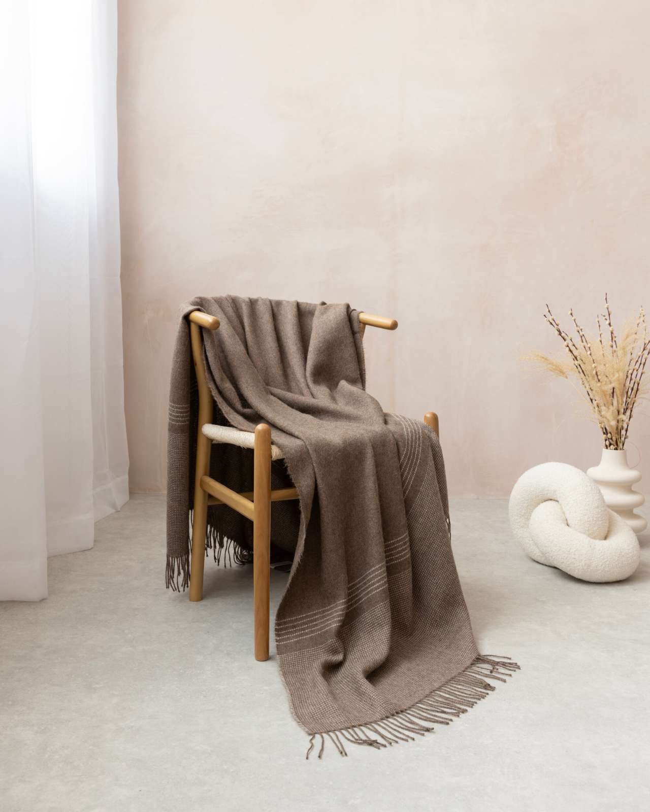 Alpaca Blanket Gift Box: Mocha Brown, featuring a woven seat chair draped with a fringed blanket, complemented by a white knot cushion and vase with dried grasses.