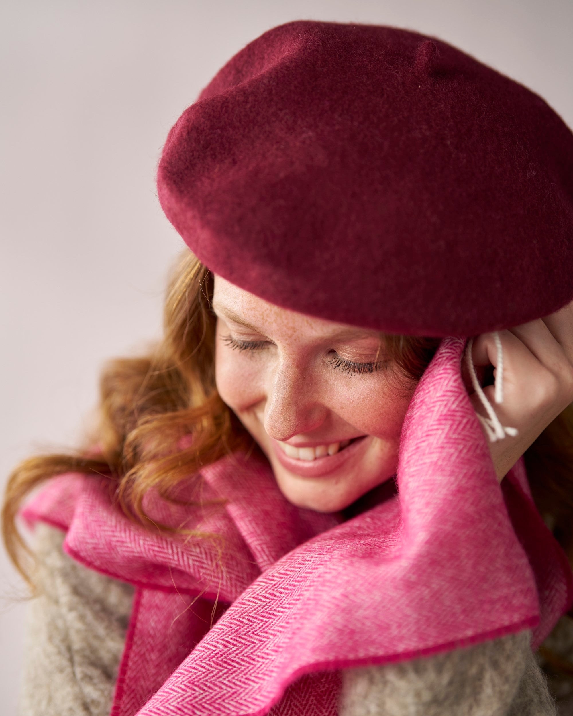 Young woman wearing a Burgundy wool beret, showcasing its thick felt texture and structured shape, paired with a bright pink herringbone-patterned scarf.