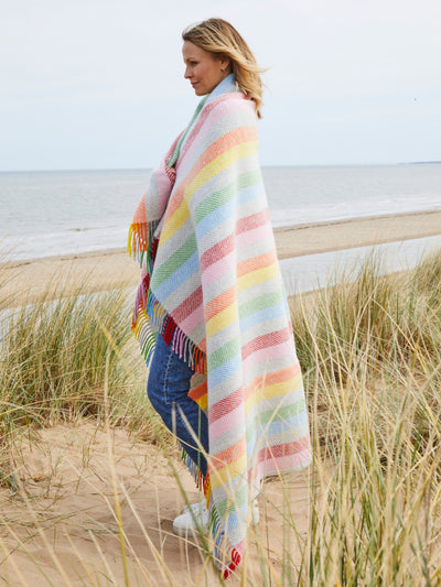 woman on a beach wrapped in rainbow stripe wool blanket from the british blanket company