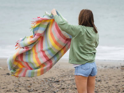 woman on a beach shaking out a rainbow stripe wool blanket from the british blanket company