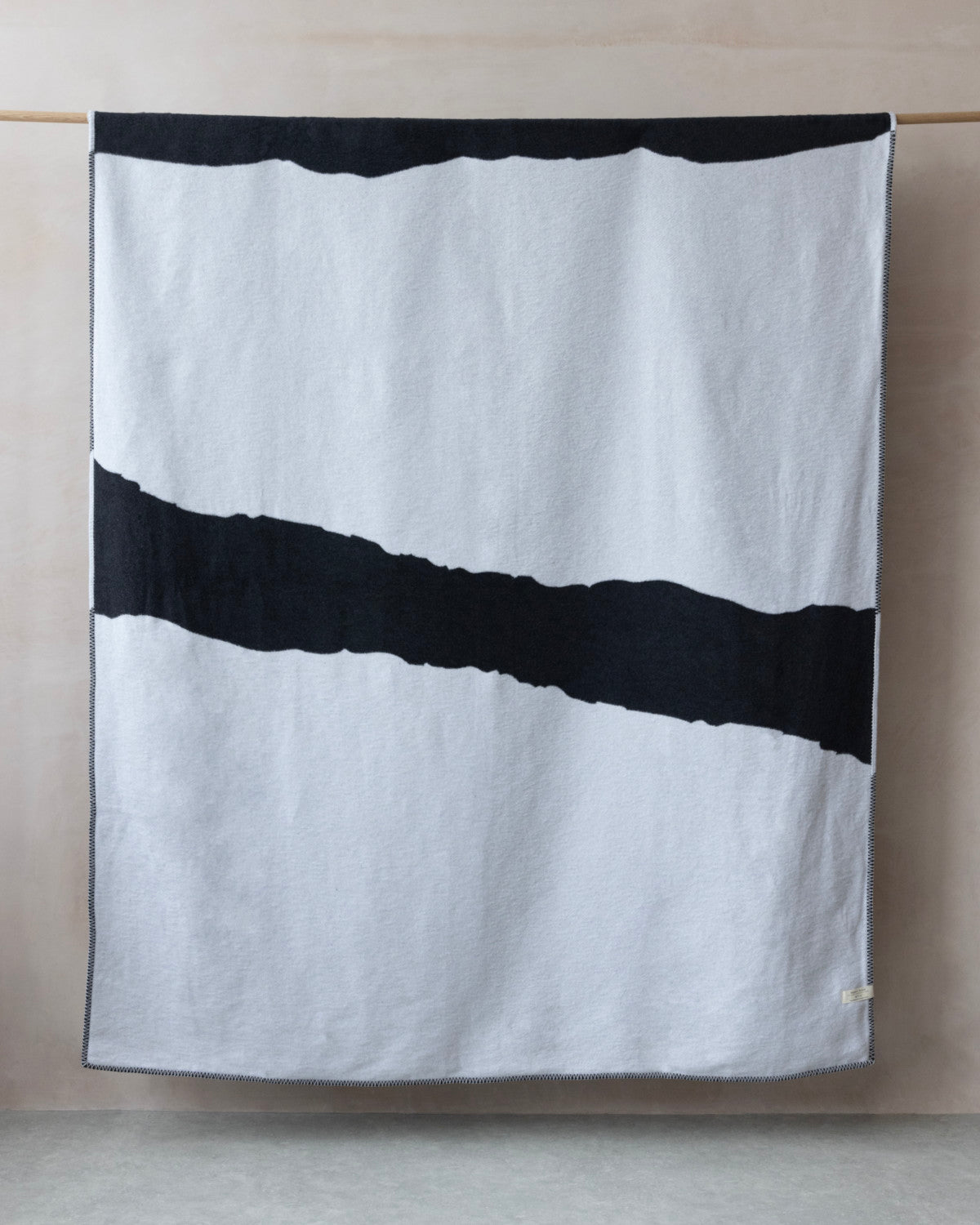 Cloud Cotton Blanket: Strata in Flint draped over a rail, featuring a broad black stripe, minimalist design, and blanket-stitched edges, crafted from recycled cotton.