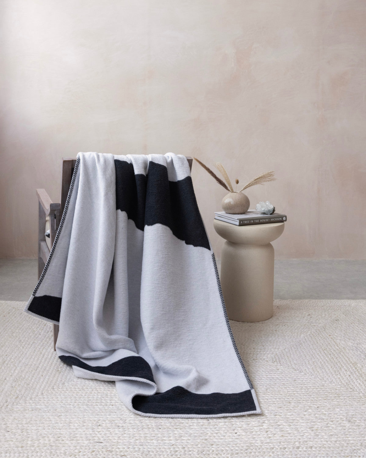 A cozy corner features the Cloud Cotton Blanket: Strata in Flint, draped over a wooden armchair, showcasing its two-tone design and soft, brushed texture.