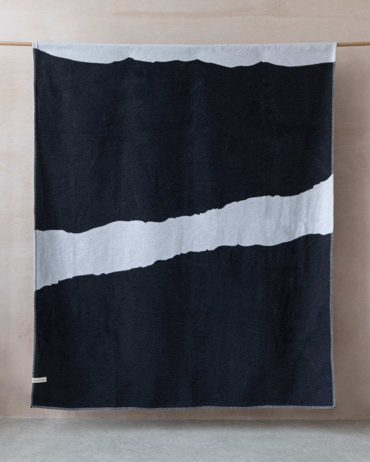Cloud Cotton Blanket: Strata in Flint, featuring a deep charcoal hue with a white torn-edge stripe and contrasting stitching, displayed on a wooden rod.