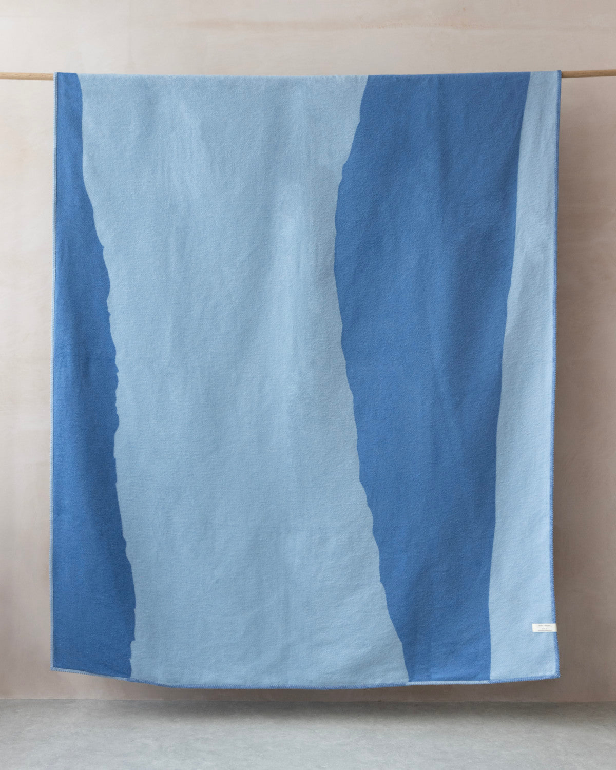 Cloud Cotton Blanket: Tide in Cornflower, featuring broad blue bands, draped over a rod. Soft, textured recycled cotton with a blanket-stitched edge.