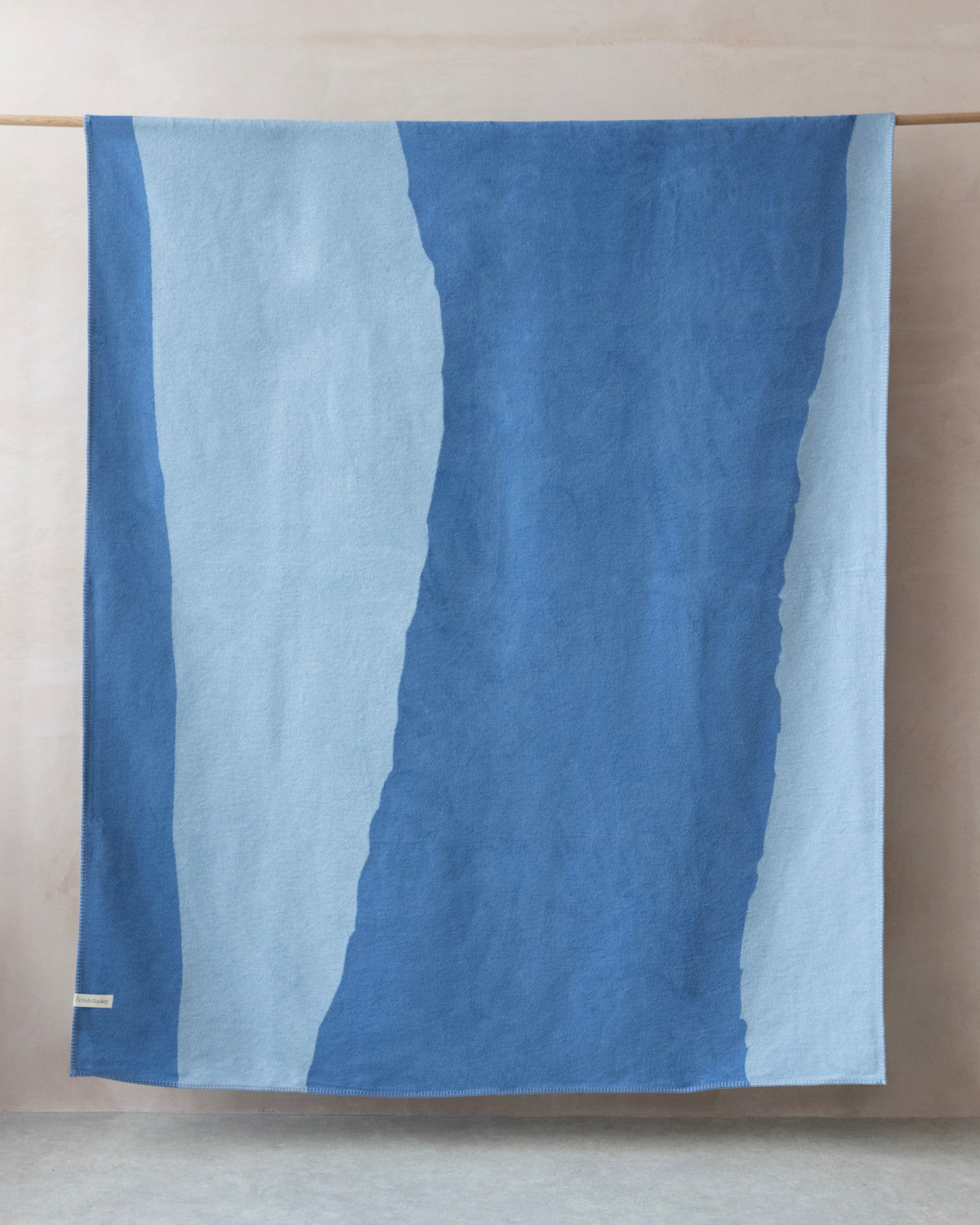The Cloud Cotton Blanket: Tide in Cornflower hangs, showcasing its abstract two-tone blue pattern and soft, fleece-like texture with a brushed finish.