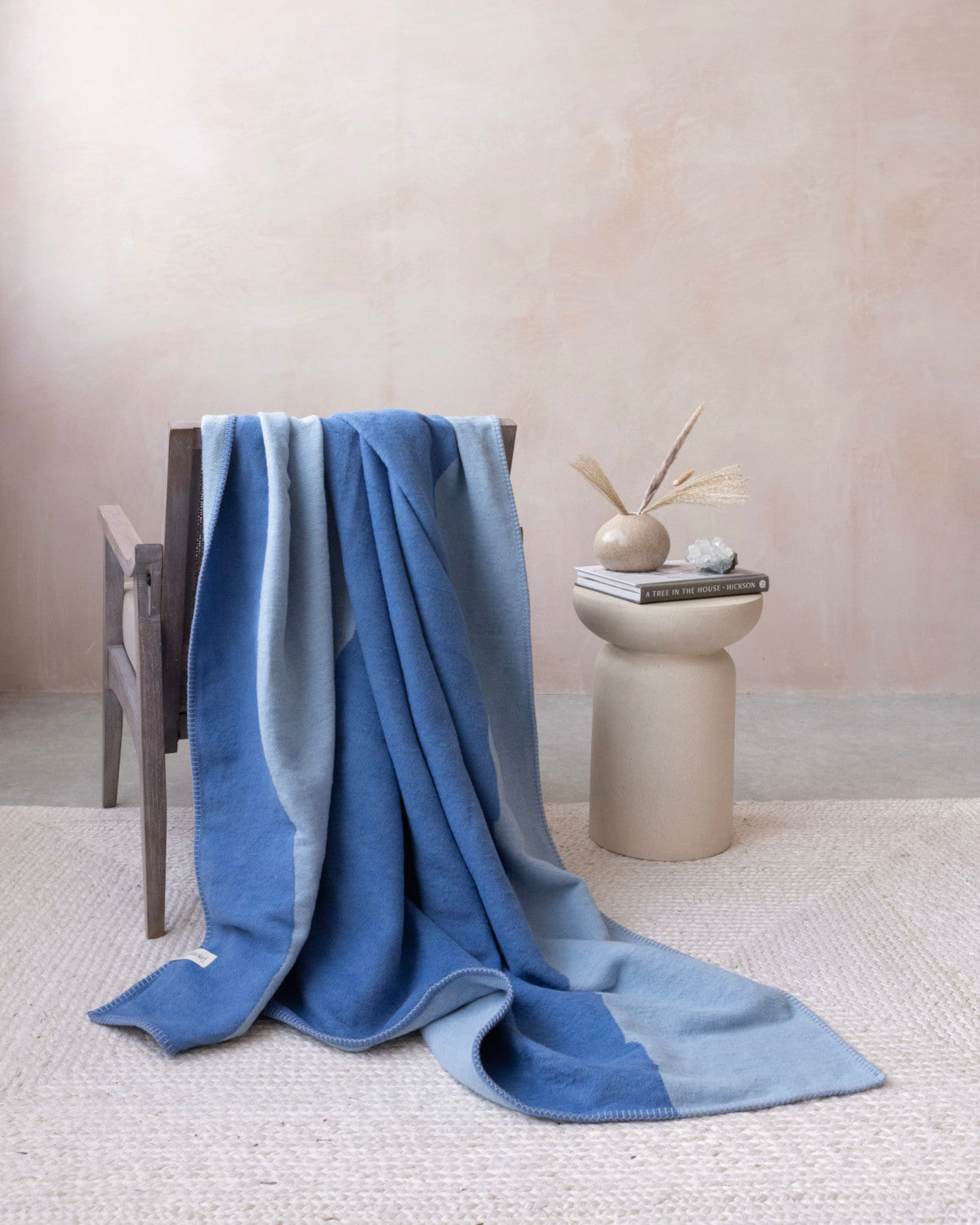 Wooden armchair with the Cloud Cotton Blanket: Tide in Cornflower, draped gracefully, showcasing its two-tone blue design, on a woven rug beside a sand-colored pedestal table.