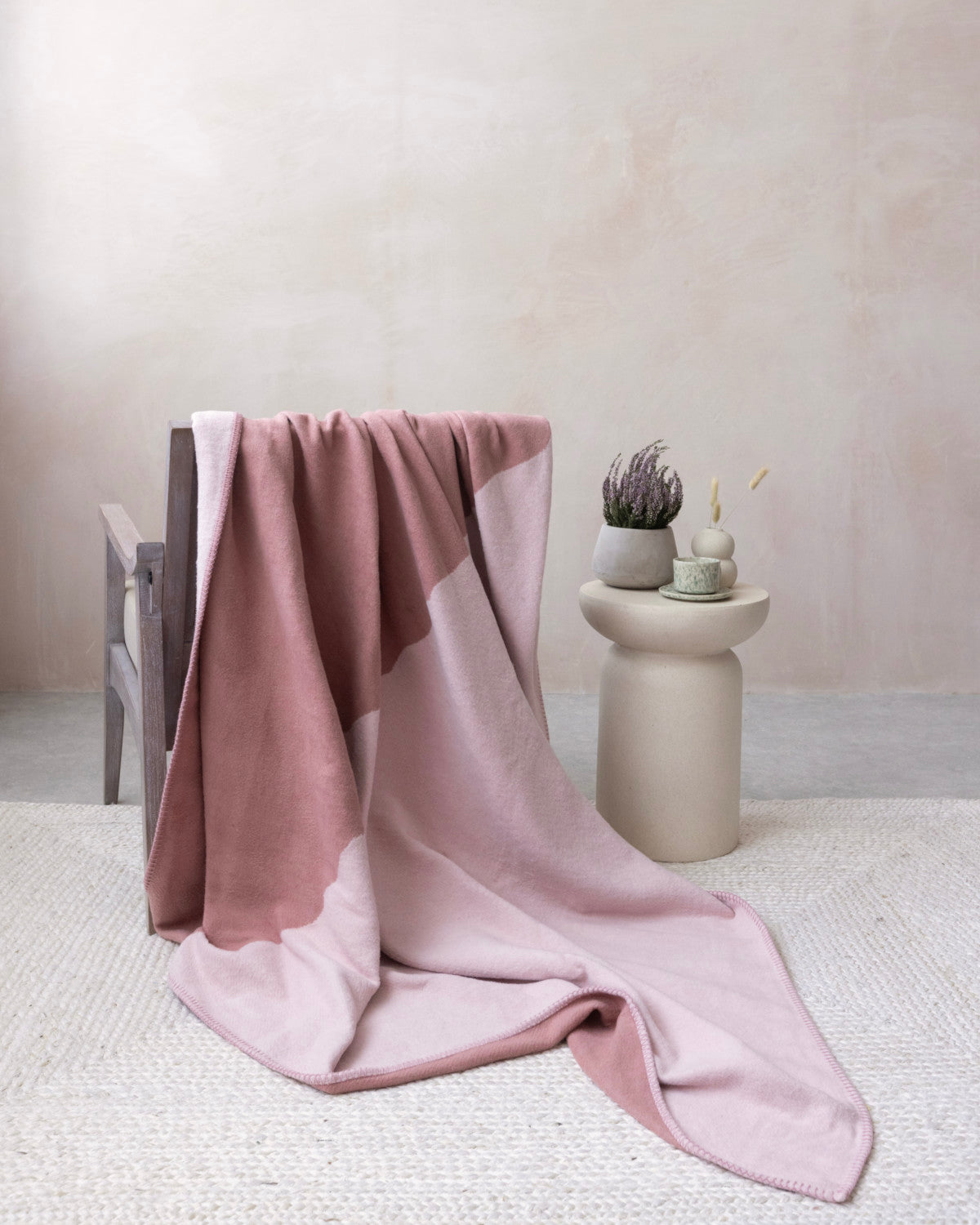 Cloud Cotton Blanket: Vale in Blush draped over a dark wood chair, showcasing its two-tone pink design and soft, brushed finish, alongside a ceramic table with decor.
