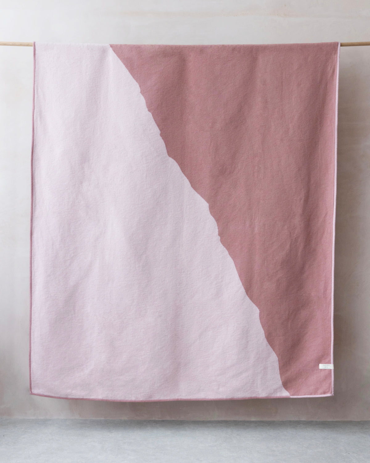 Cloud Cotton Blanket: Vale in Blush draped over a wooden rod, showcasing its soft texture and diagonal two-tone design with a visible tag.