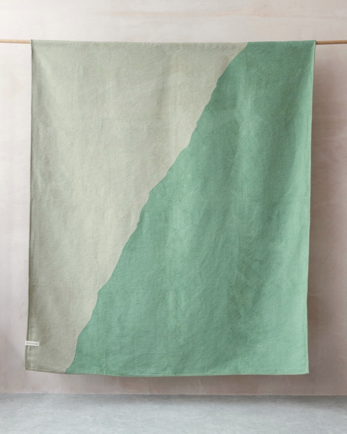 Cloud Cotton Blanket: Vale in Thyme draped over a wooden rod, featuring a two-tone design with a diagonal seam, showcasing its soft, cozy texture.