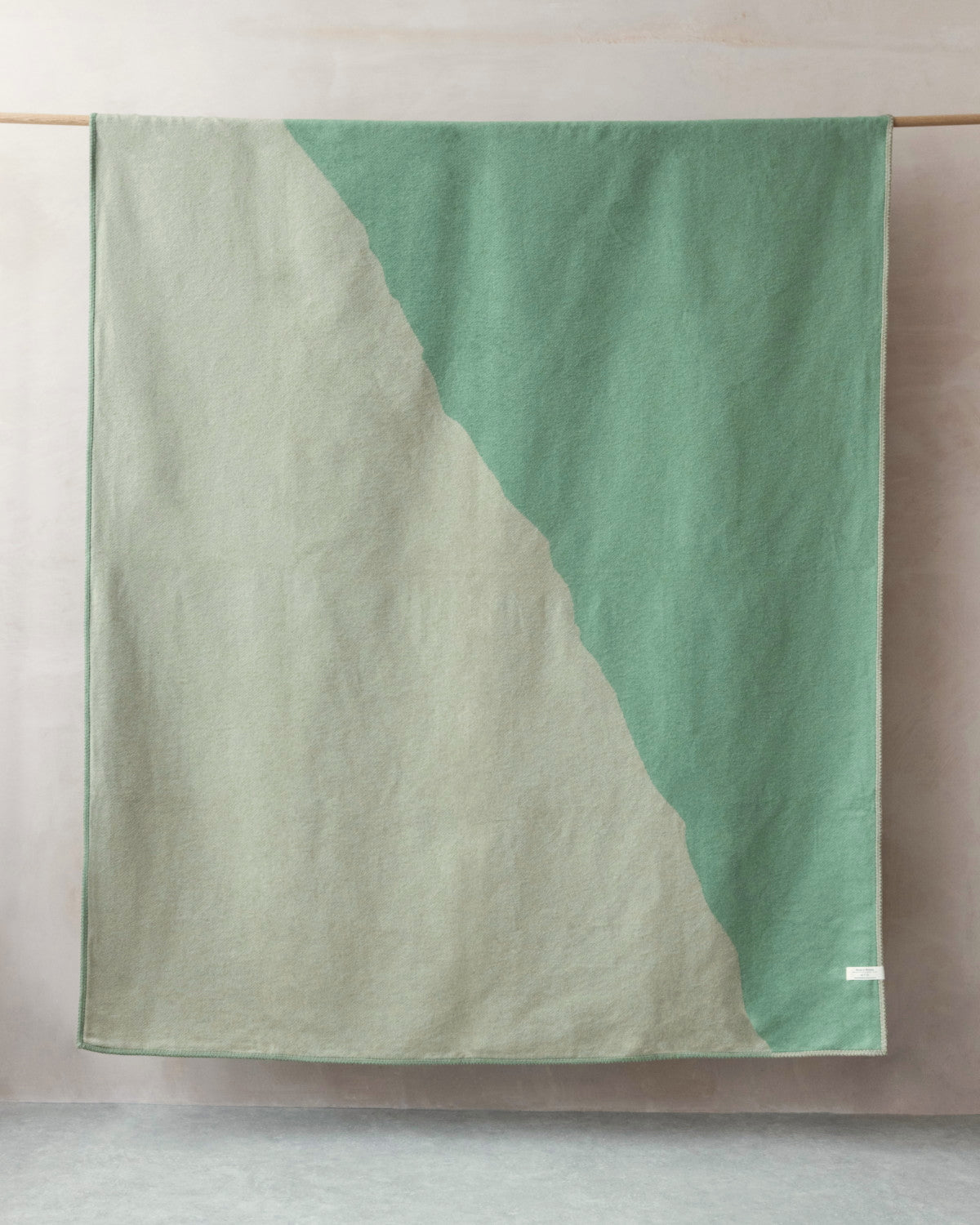 Cloud Cotton Blanket: Vale in Thyme draped on a rod, showcasing its two-tone design with minty-green and stone-gray sections, finished with a contrasting stitch.