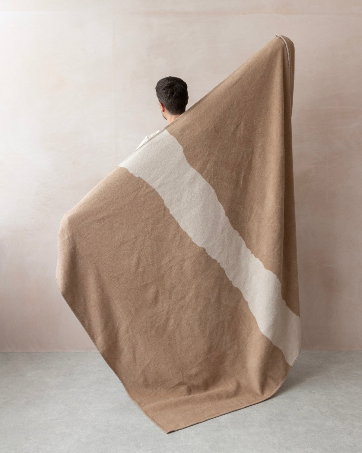 Person holds Cloud Cotton Blanket: Strata in Clay, featuring a warm camel-tan color with off-white diagonal stripe, showcasing its soft, brushed texture.