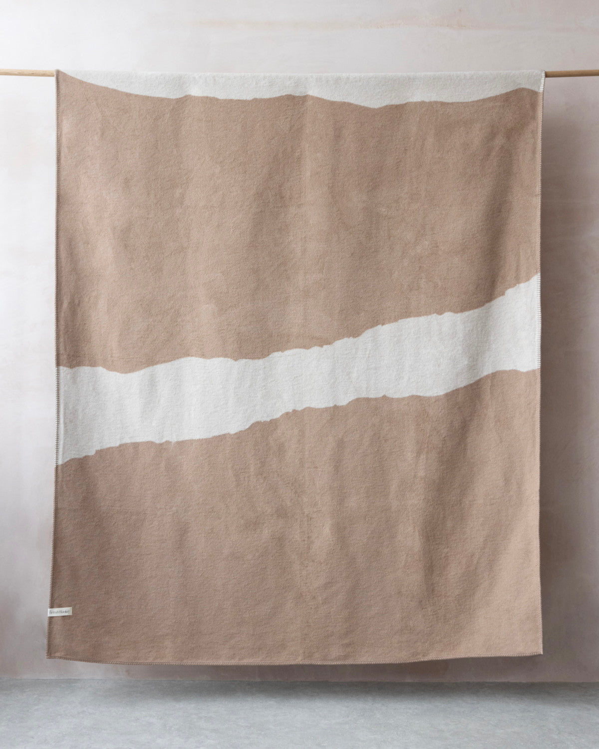 Cloud Cotton Blanket: Strata in Clay, featuring a muted tan hue with a white band, draped on a wooden rod, showcasing its soft texture and blanket-stitched edge.