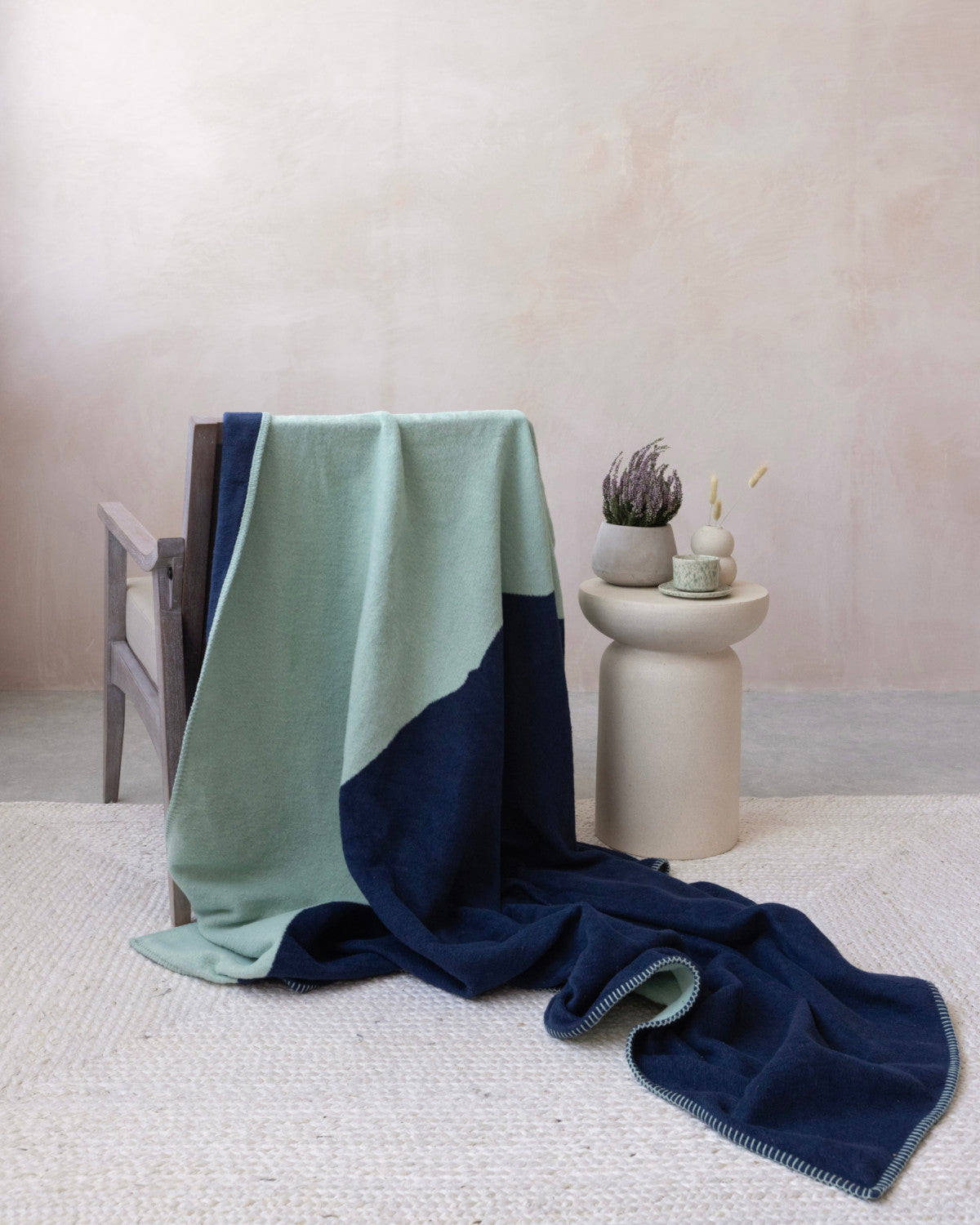 Wooden-frame armchair with Cloud Cotton Blanket: Vale in Mistral, mint and navy, on a beige side table with decor, creating a minimalist, serene setting.