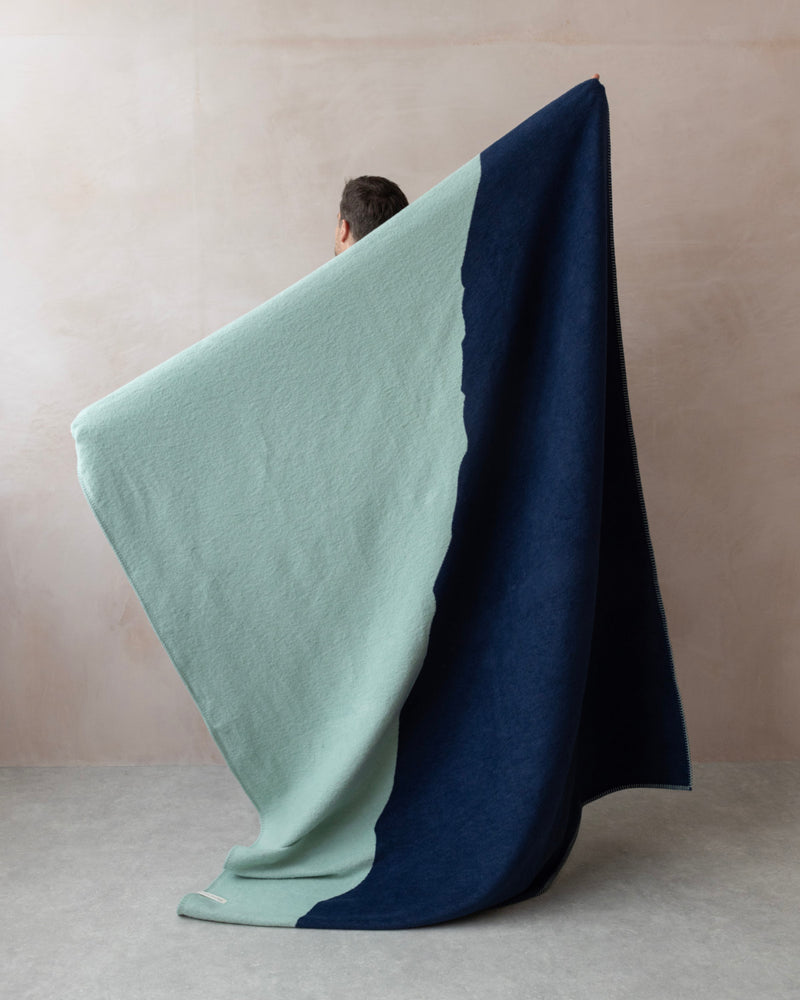 Person holds the Cloud Cotton Blanket: Vale in Mistral, showcasing its two-tone mint-green and navy design, highlighting its soft, brushed finish against a neutral background.