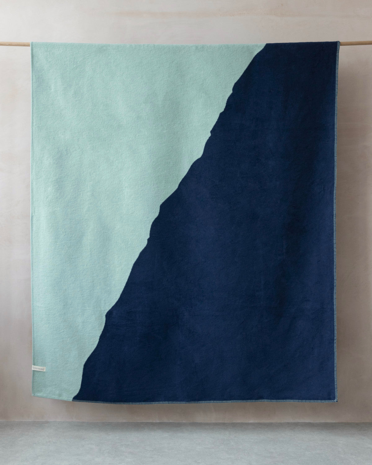 Cloud Cotton Blanket: Vale in Mistral, featuring a two-tone design with mint-green and navy, hangs neatly from a dowel, showcasing its mountain-like diagonal division.
