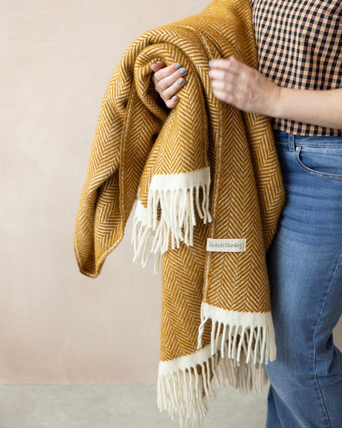 Clifton Herringbone Blanket: Spiced Cider draped over an arm, showcasing its mustard-yellow wool texture with off-white fringed edges and a British Blanket label.