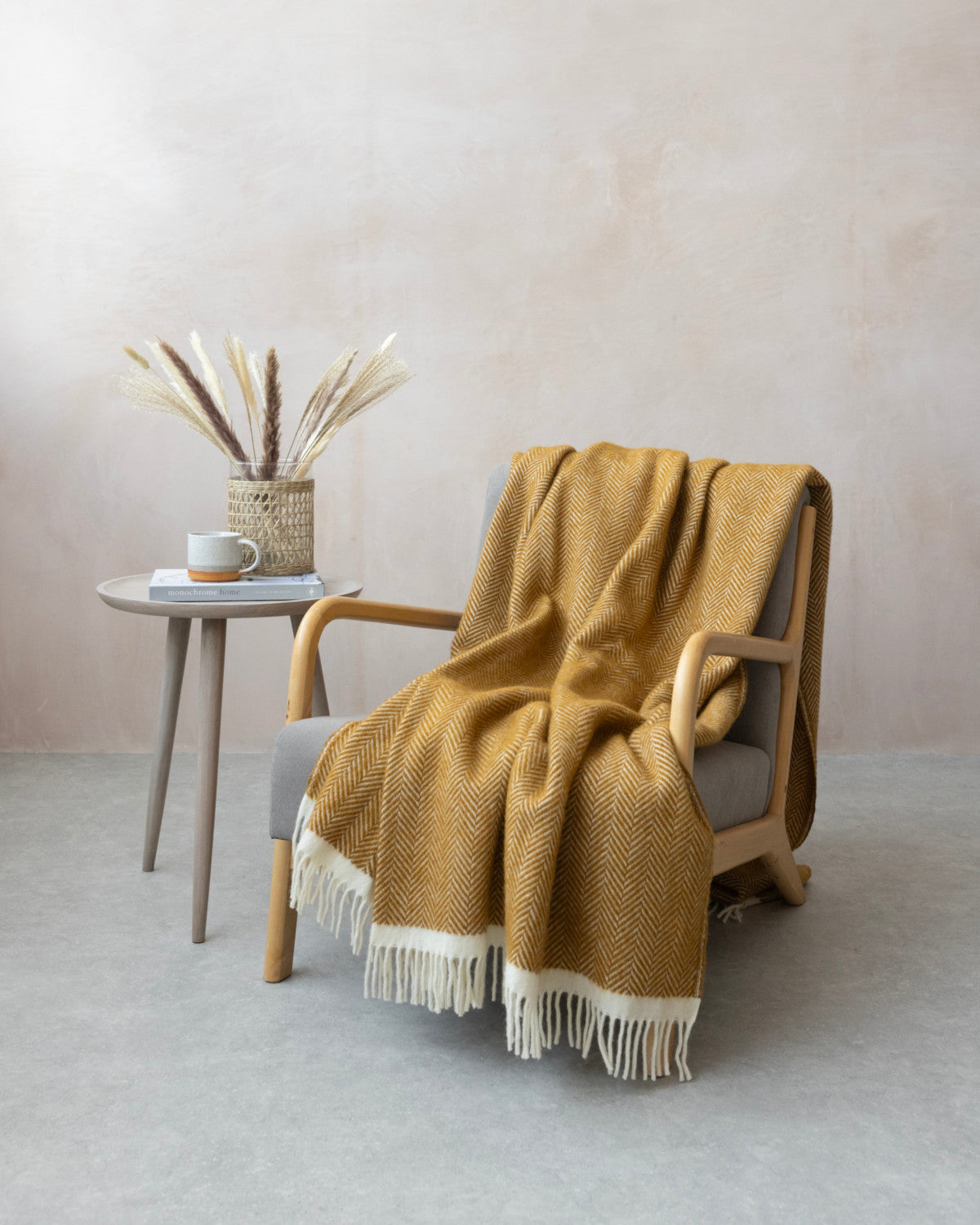 Cozy corner featuring a lounge chair with Clifton Herringbone Blanket: Spiced Cider draped over, alongside a side table with a book, mug, and dried grasses.