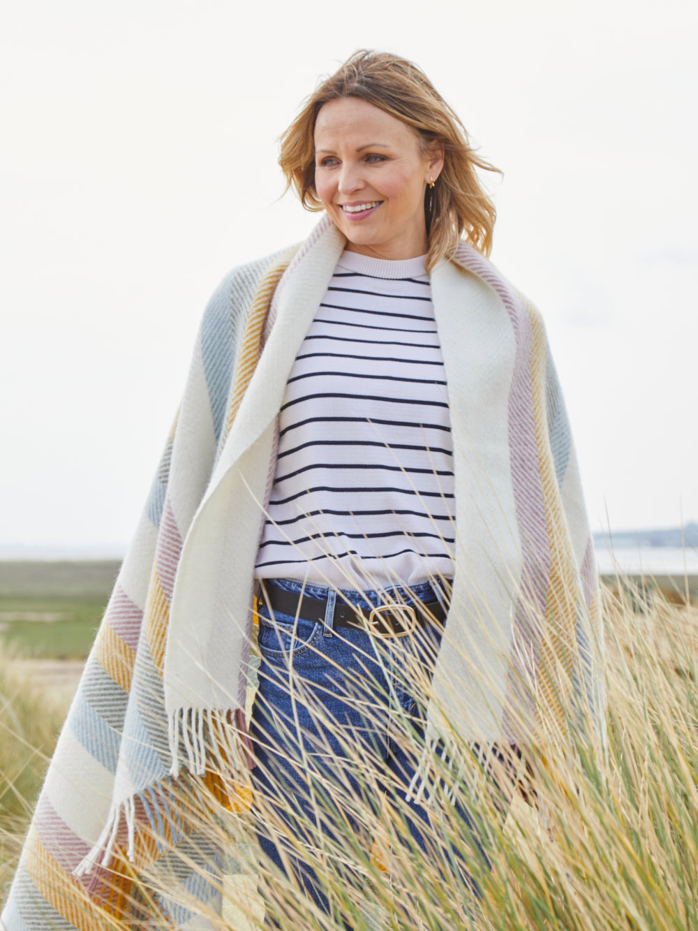 Woman on the beach wrapped in Pastel Rainbow Stripe Herringbone Throw by The British Blanket Company