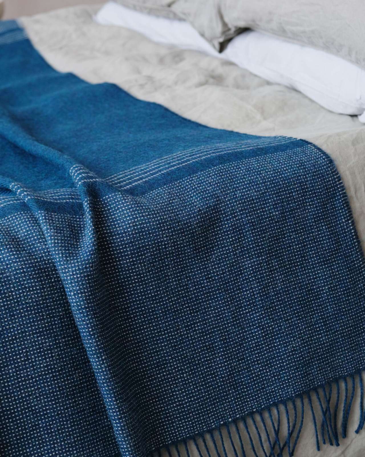 Teal Blue Alpaca Blanket drapes a bed with neutral bedding, featuring a dotted weave and fringe, part of the thoughtful Alpaca Blanket Gift Box.