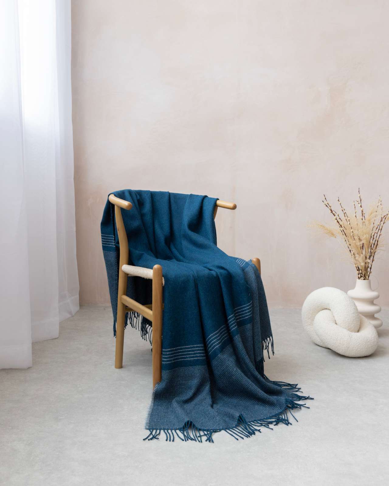 Alpaca Blanket Gift Box in Teal Blue draped over a wooden chair, with a vase of dried grasses and a sculptural knot cushion nearby.
