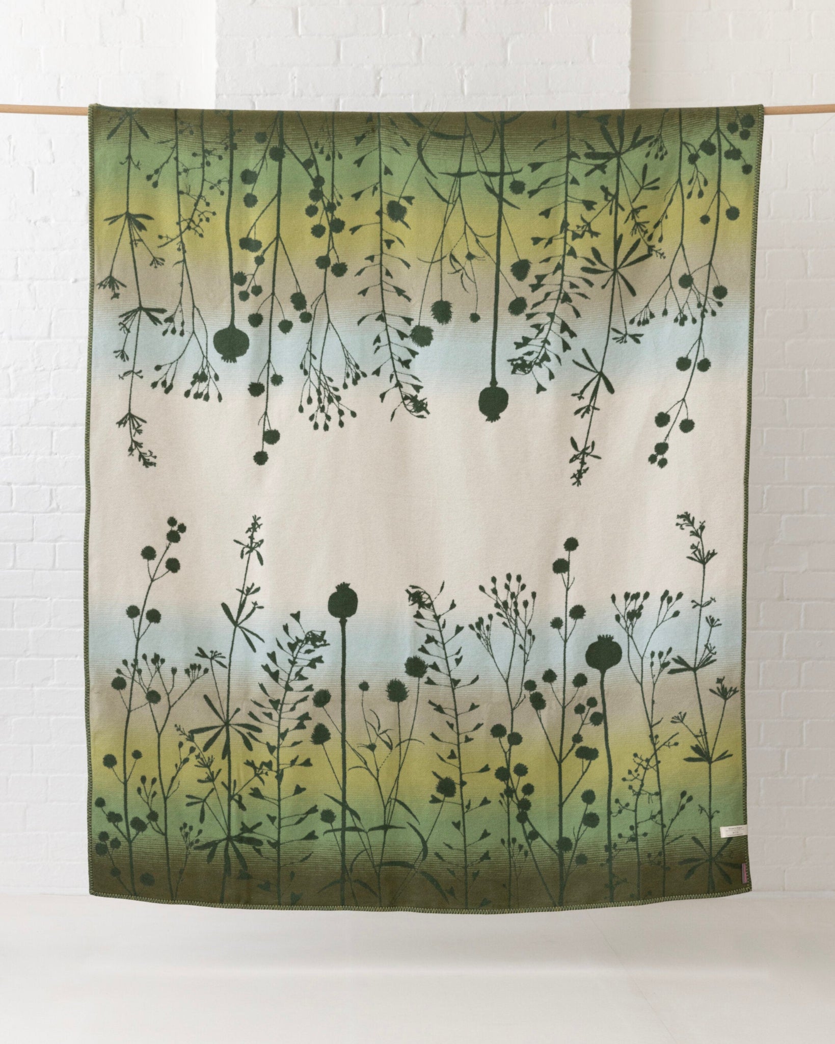 Clarissa Hulse Blanket: Seedheads in Moss, a large jacquard-woven cotton throw with a gradient of greens and dark wildflower silhouettes, hanging on a wooden rod.