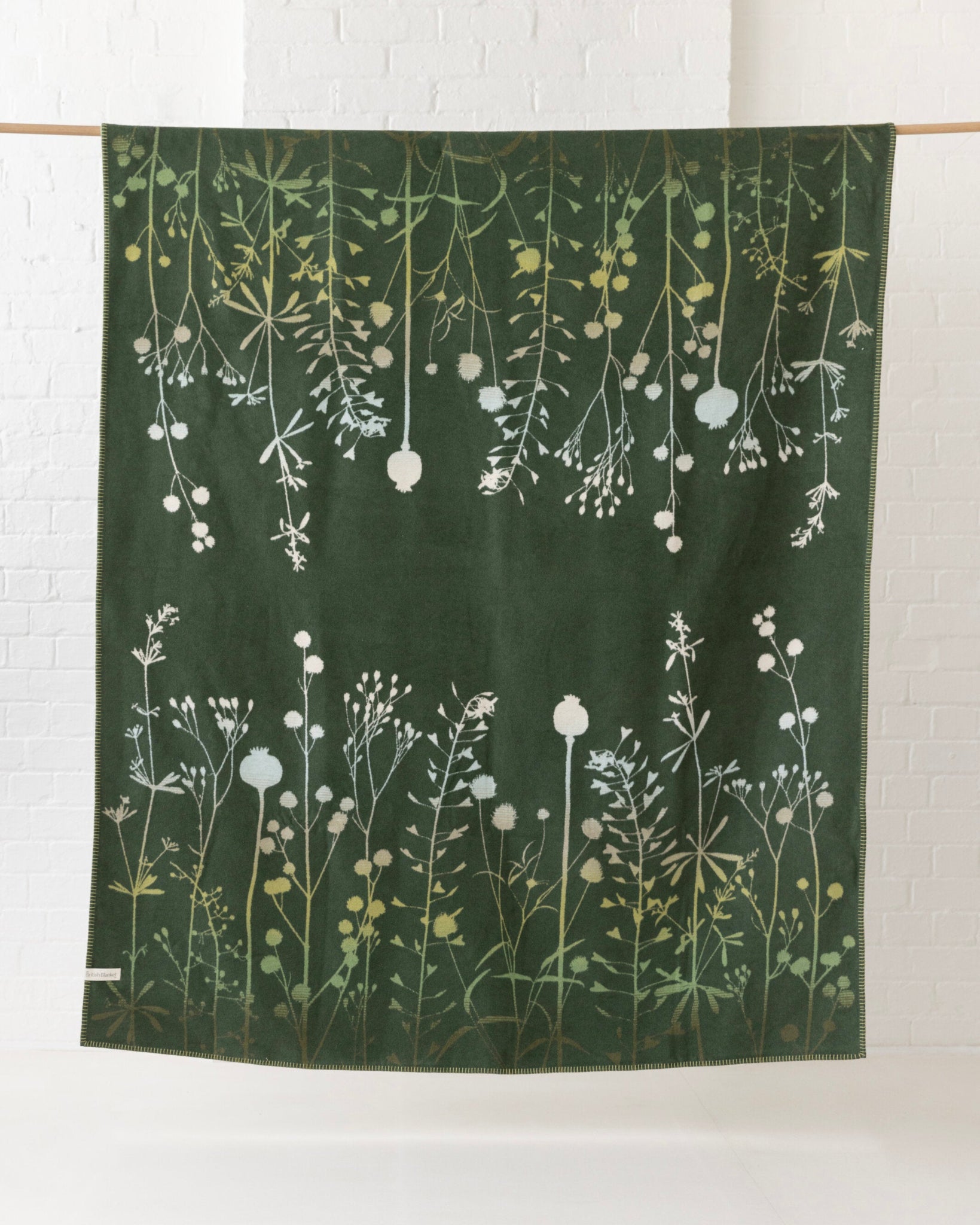 Clarissa Hulse Blanket: Seedheads in Moss, a large green jacquard-woven throw with wildflower motifs, hangs elegantly against a white brick wall.