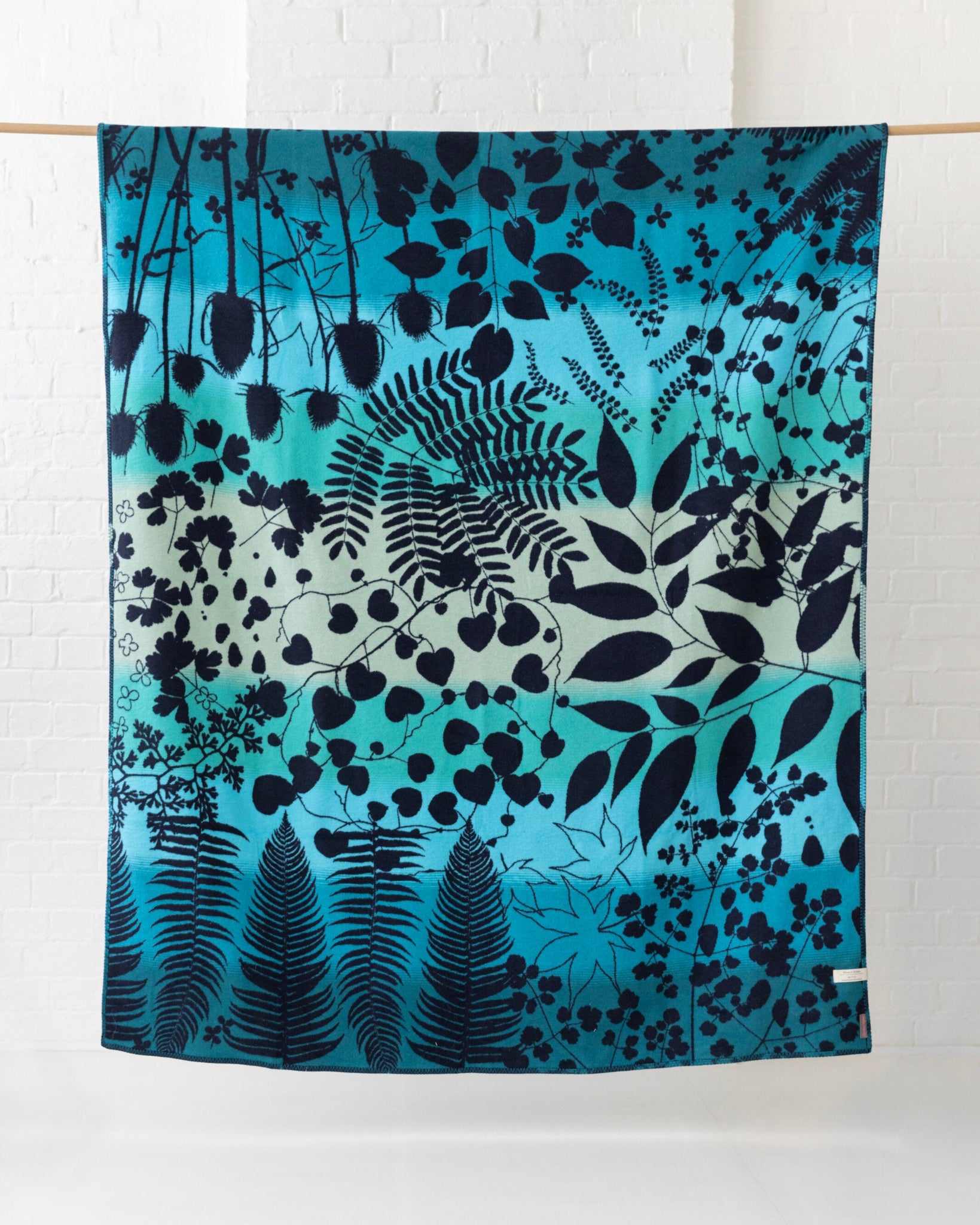 Clarissa Hulse Blanket: Indian Summer in Peacock, featuring teal gradient cotton with black botanical silhouettes, woven on jacquard looms, displayed against a white brick wall.