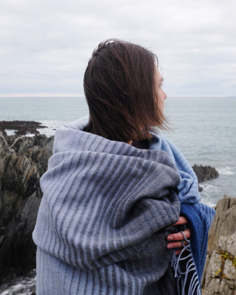 Person stands on rocky shore wrapped in Shipping Forecast 'Ocean Ombre' Merino Blanket, gazing at ocean, showcasing its soft, striped design and craftsmanship.