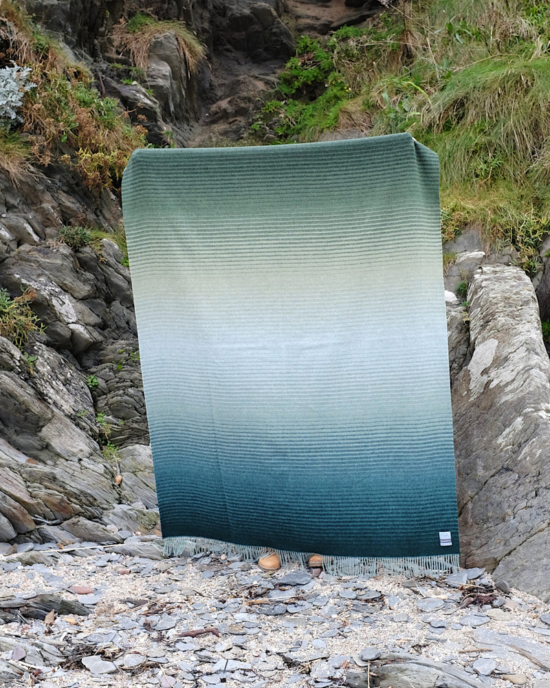 Shipping Forecast 'Coast Ombre' Merino Blanket held by shoreline, showcasing its ombré texture from deep teal to darker green, crafted in Britain from 100% Merino lambswool.