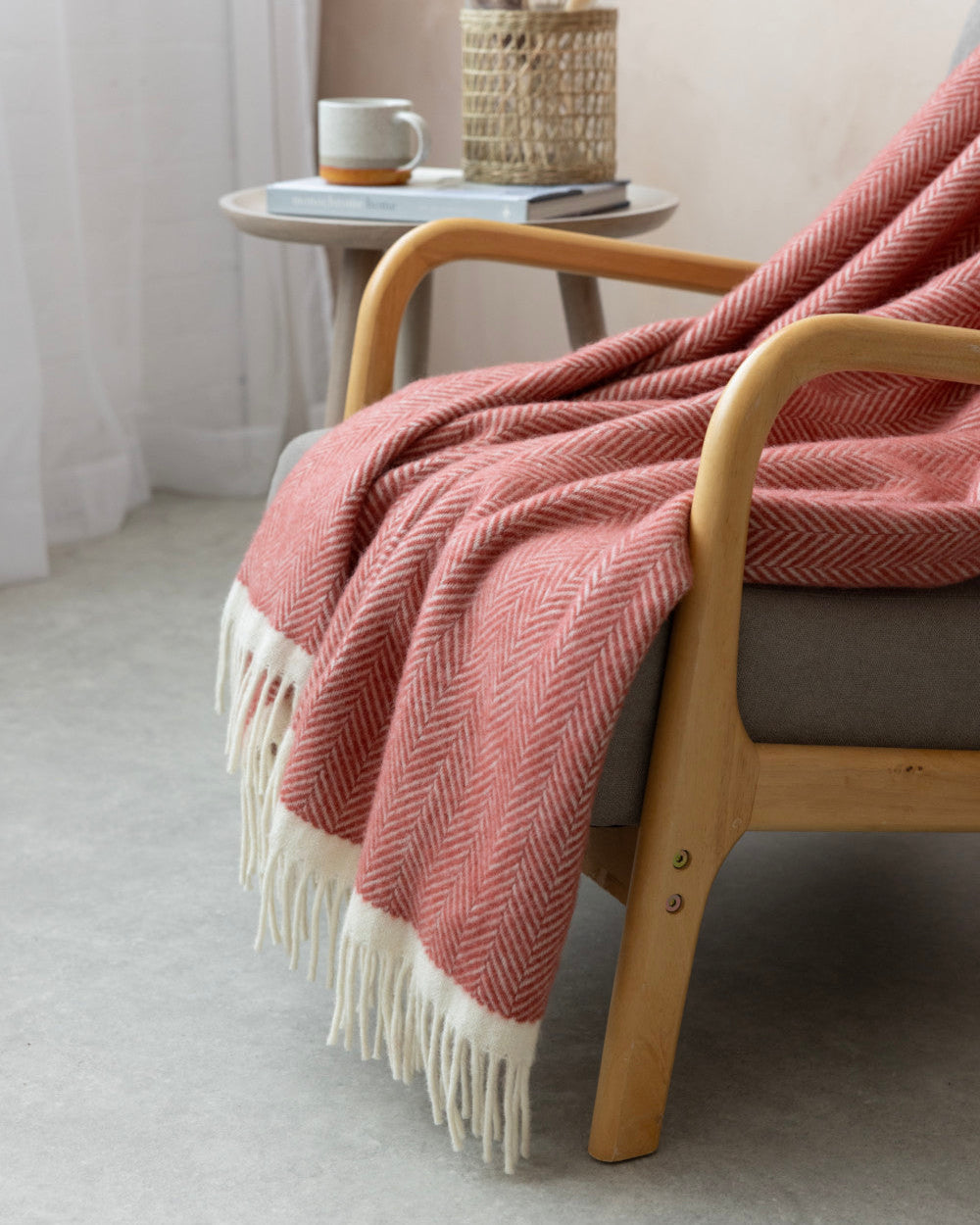 Cozy scene showcasing Clifton Blanket Gift Box: Rose, featuring a herringbone-patterned wool blanket on an armchair, alongside a side table with a mug and books.