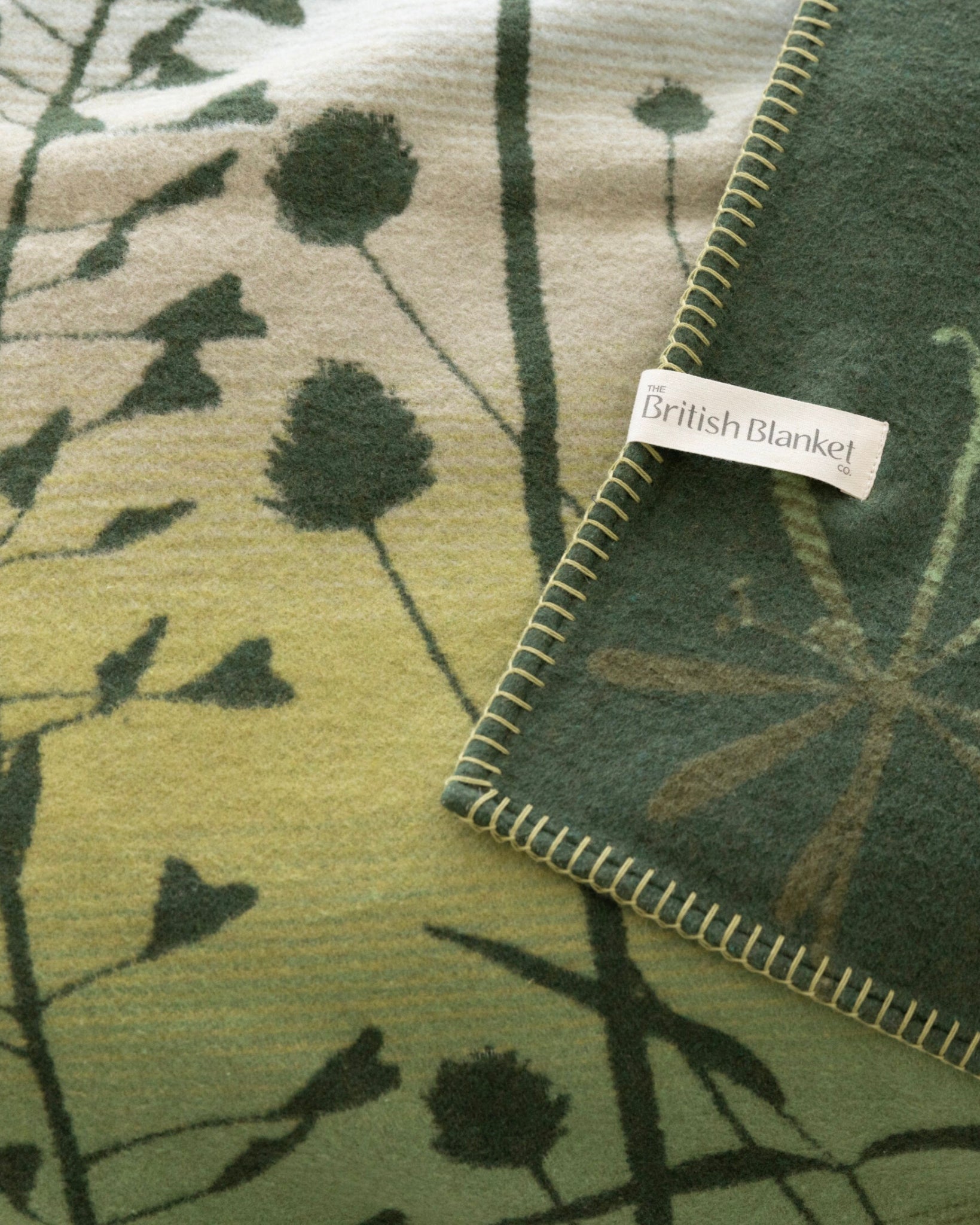 Clarissa Hulse Blanket: Seedheads in Moss features a two-tone green and cream design with botanical silhouettes, woven label, and whip-stitched edges for a soft, natural look.
