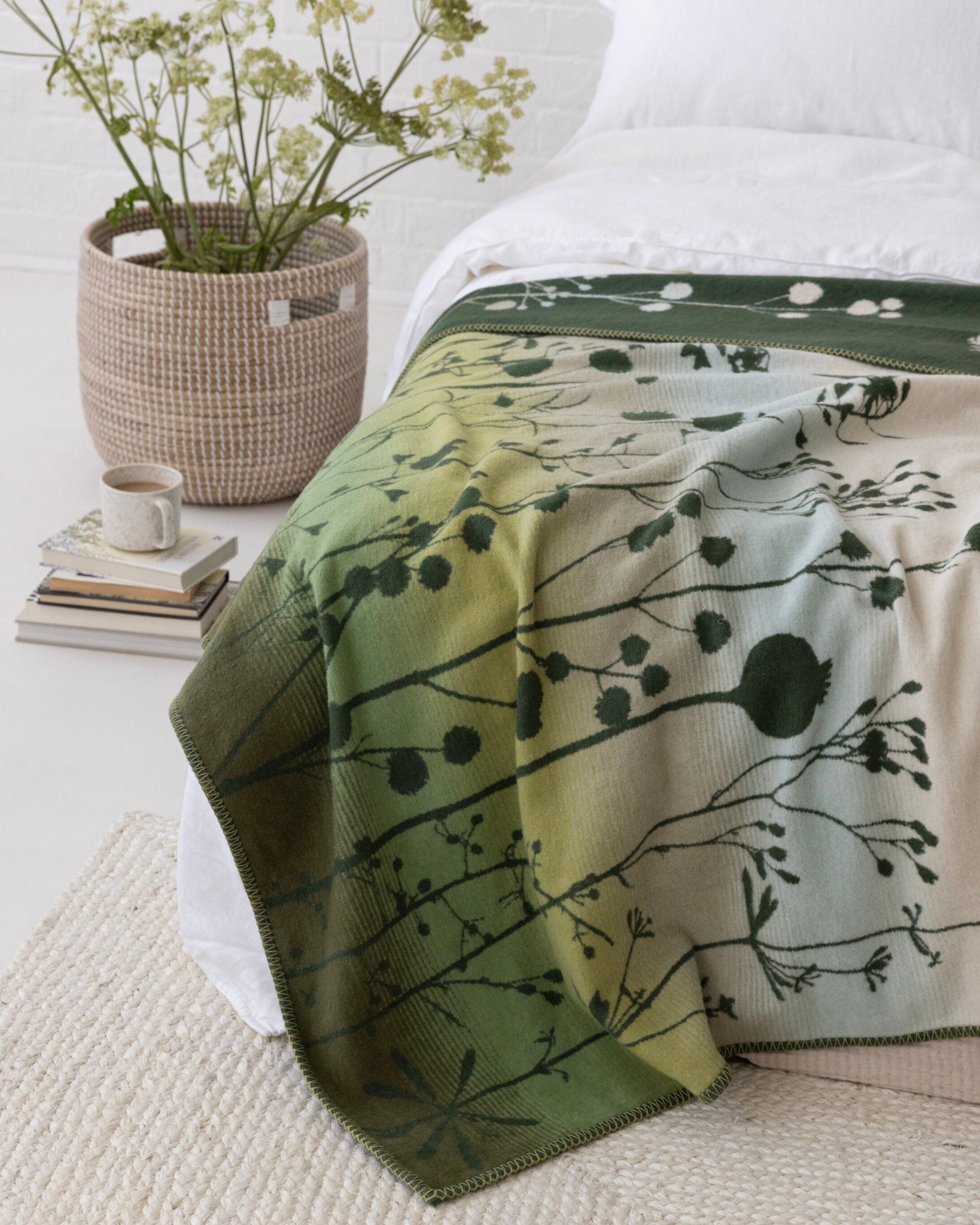 Clarissa Hulse Blanket: Seedheads in Moss drapes over a bed, showcasing its green ombré and botanical pattern. Nearby, books, a mug, and wildflowers enhance the serene setting.