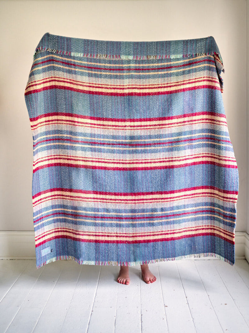Random recycled wool blanket (C) draped with fringed edges, revealing bare feet on a white floor, showcasing its hand-woven design with broad horizontal bands.