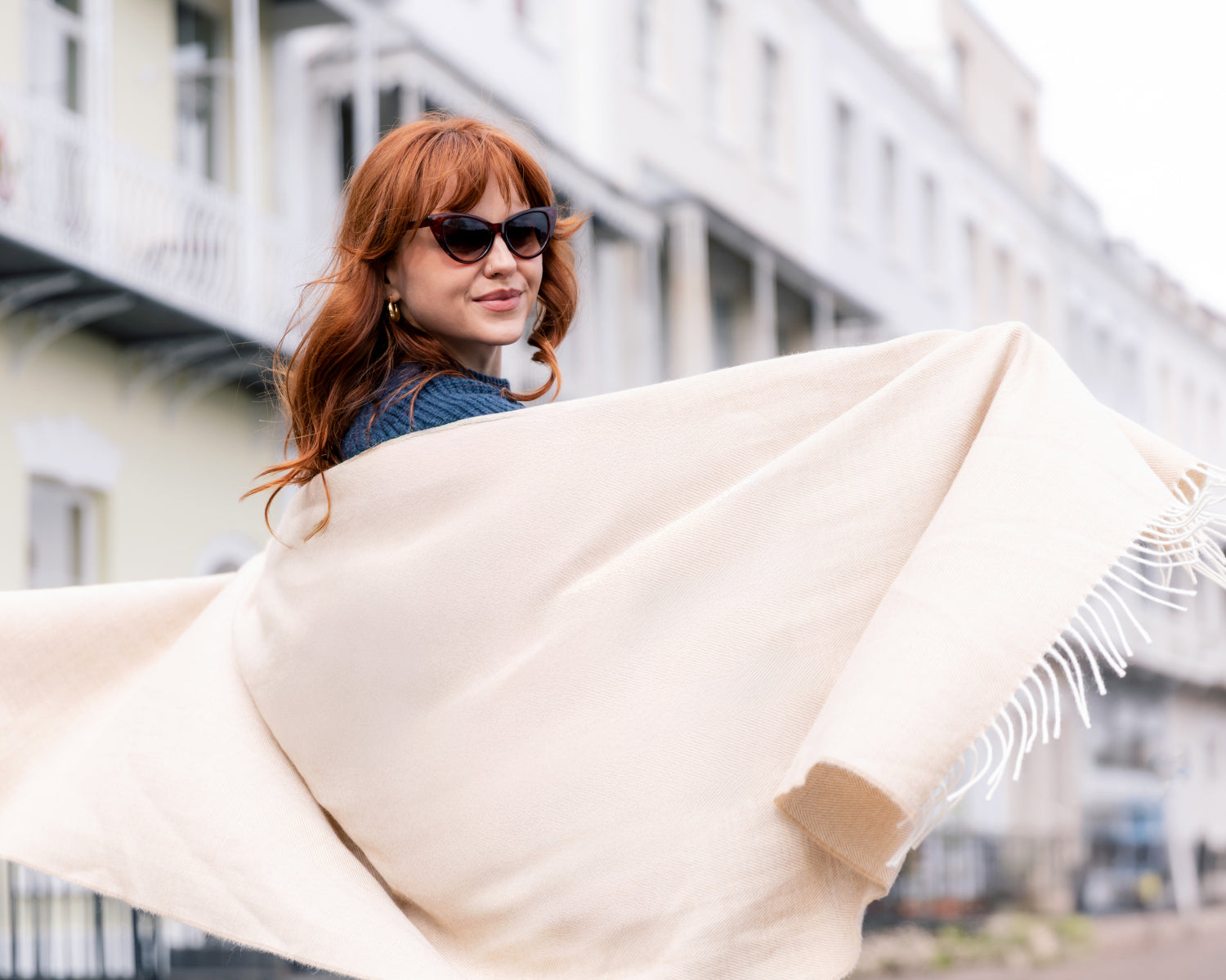 Woman wearing the Oversized Blanket Scarf in Basswood Herringbone, billowing behind her, with sunglasses and gold hoop earrings, showcasing the scarf's elegance and versatility.