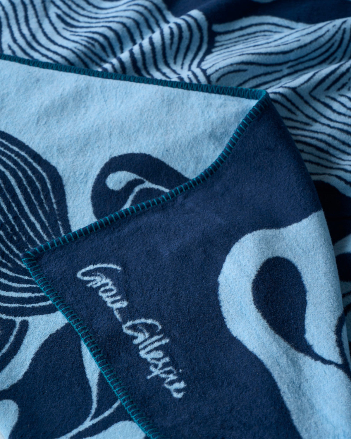 Close-up of the Cloud Cotton Blanket: Blue Prayer Plant, showcasing its wavy blue patterns, teal overlock stitching, and cursive branding, woven from sustainable cotton.