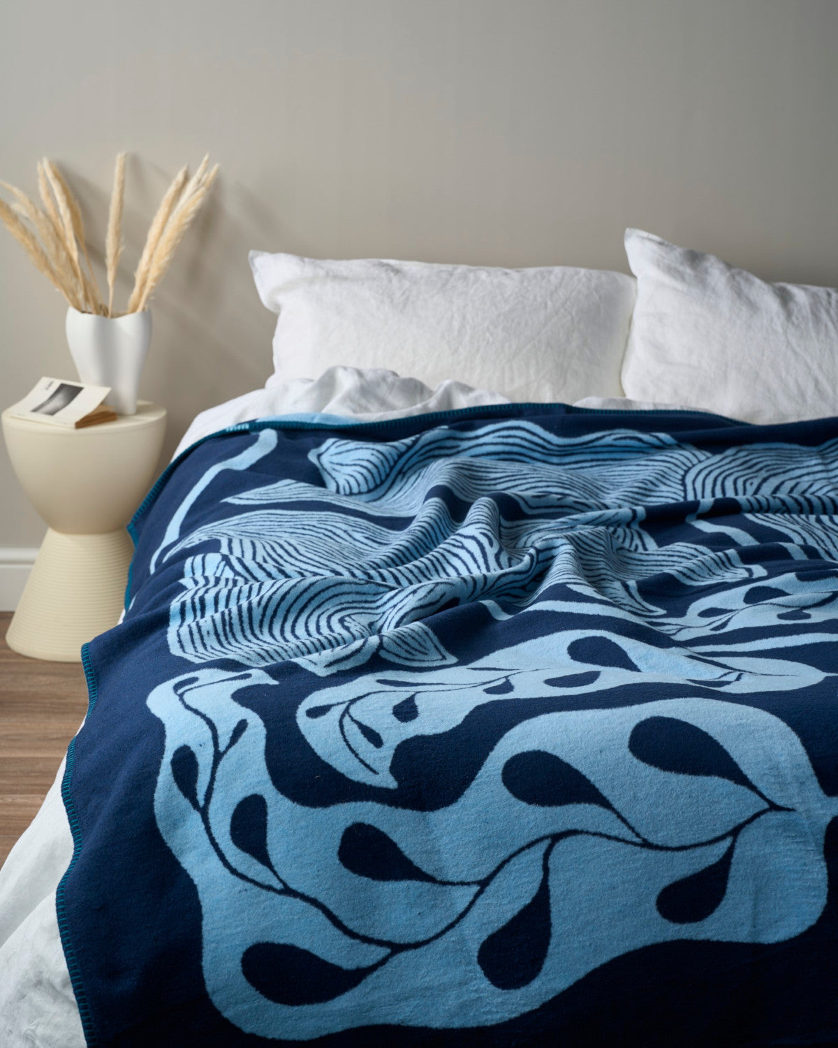 The Cloud Cotton Blanket: Blue Prayer Plant (C) with botanical design drapes over a bed, accompanied by a side table with pampas grass and an open book.