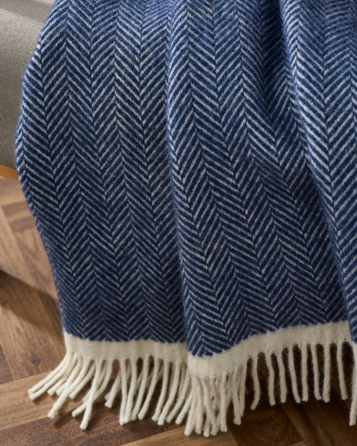 Clifton Blanket Hamper: Brunel displayed with a blue-and-off-white herringbone design, cream band, and fringes, elegantly draped over furniture on parquet flooring.