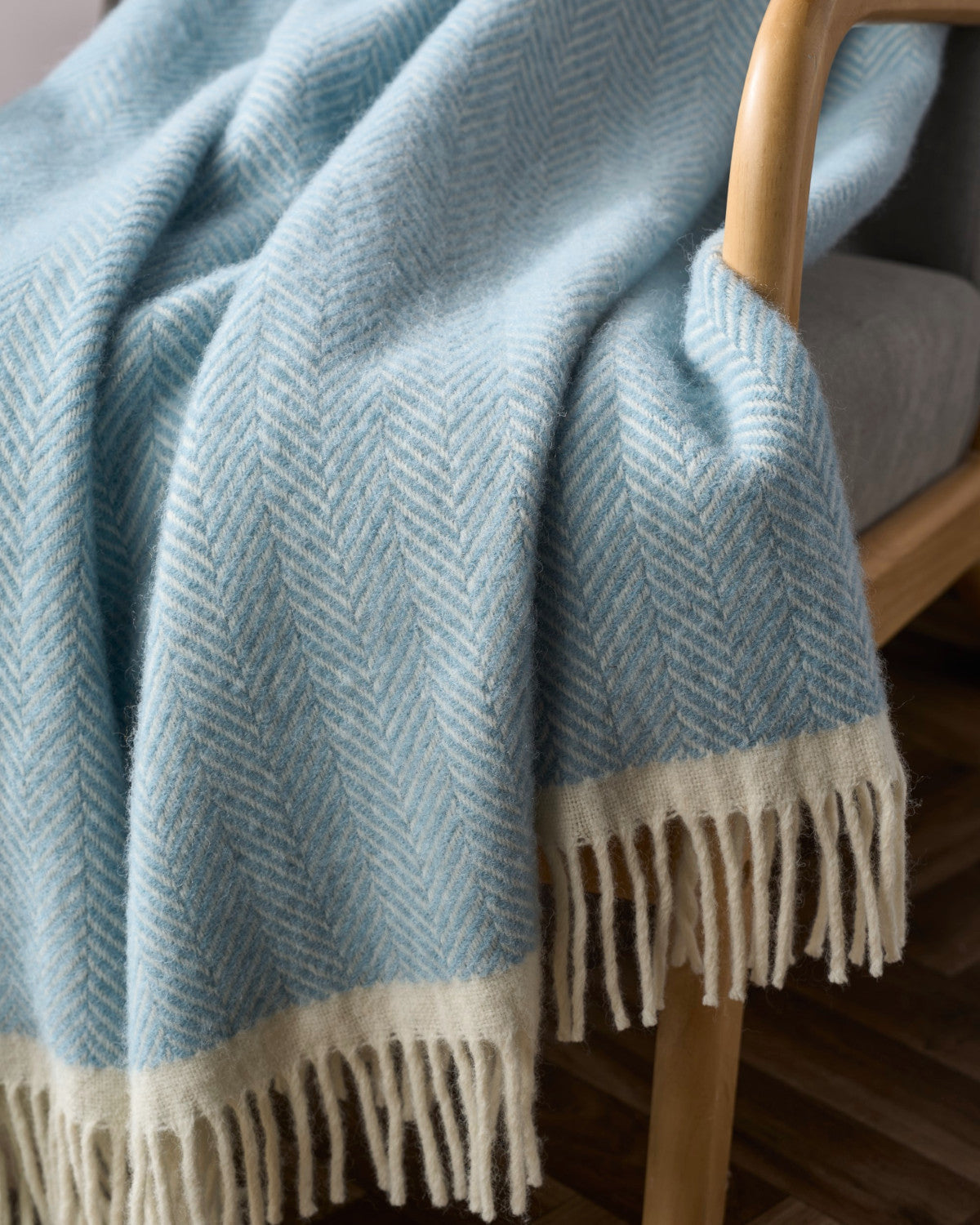 Clifton Blanket Hamper: Harbourside, a soft woolen throw with a herringbone pattern, creamy-white edges, and white fringe, draped over a wooden chair.
